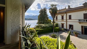 A balcony with potted plants, a view of a lake, and a building with a traditional architectural style. - 2 bedroom awesome home in Orta San Giulio NO (Orta San Giulio NO)
