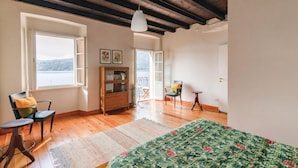 A room with wooden floors, a bed with a floral bedspread, a chair with a cushion, a small table, a cabinet, and a view of a body of water through a window. - 2 bedroom awesome home in Orta San Giulio NO (Orta San Giulio NO)