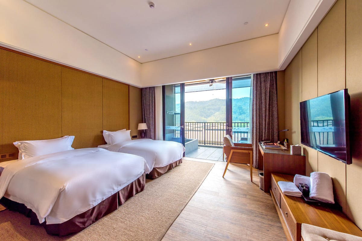 Quarto de hotel com duas camas, uma escrivaninha, televisão e varanda com vista para as montanhas.