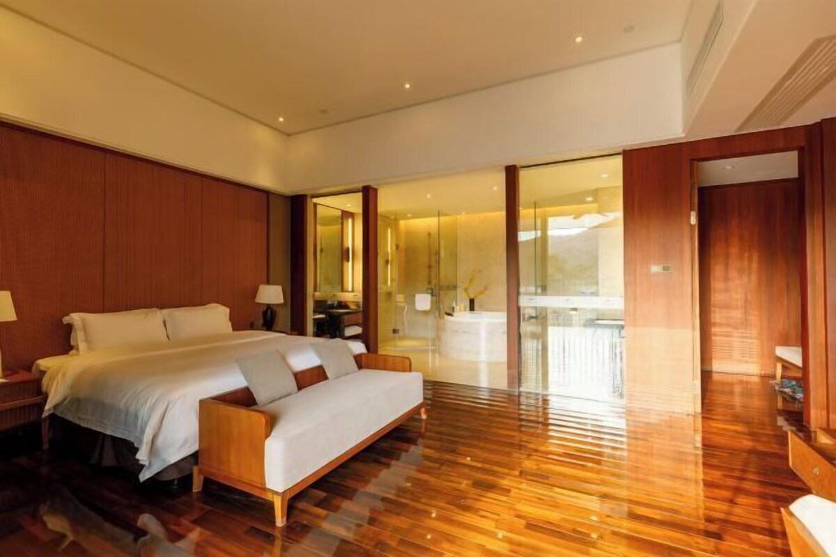 A modern hotel room with a wooden floor, a large bed with white linens, a sofa, a glass shower enclosure, and a wooden wardrobe.