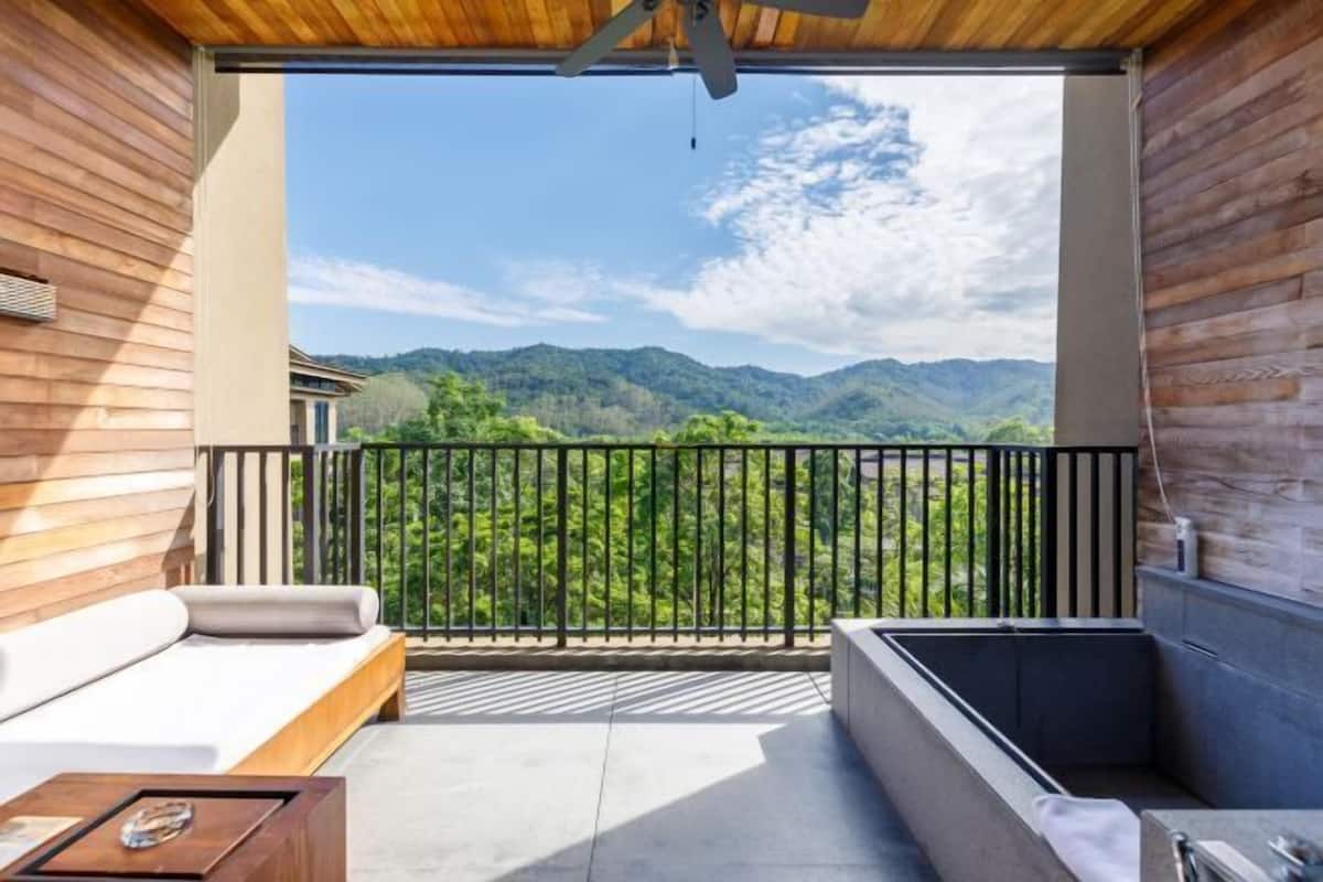 Varanda com piso de madeira, um sofá, uma mesa pequena e vista para montanhas verdes.
