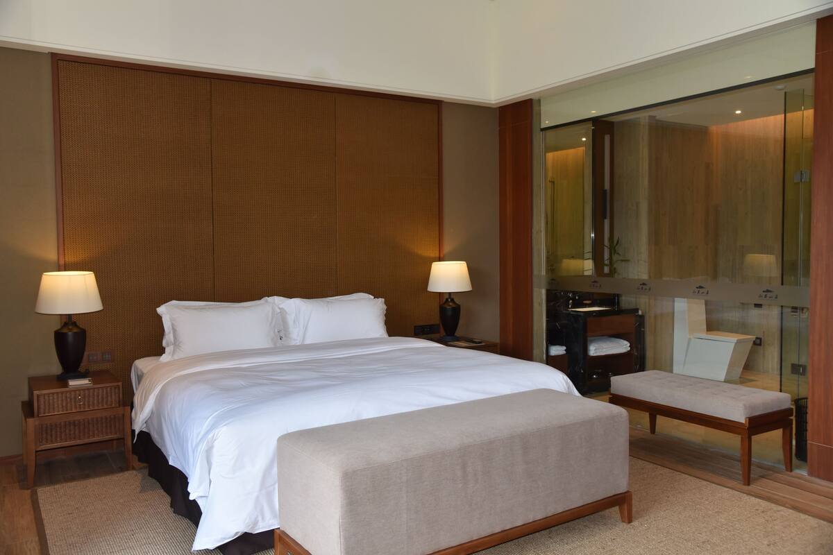 Quarto de hotel moderno com uma cama grande, duas luminárias de cabeceira, um banco, um espelho e um box de vidro para o chuveiro.