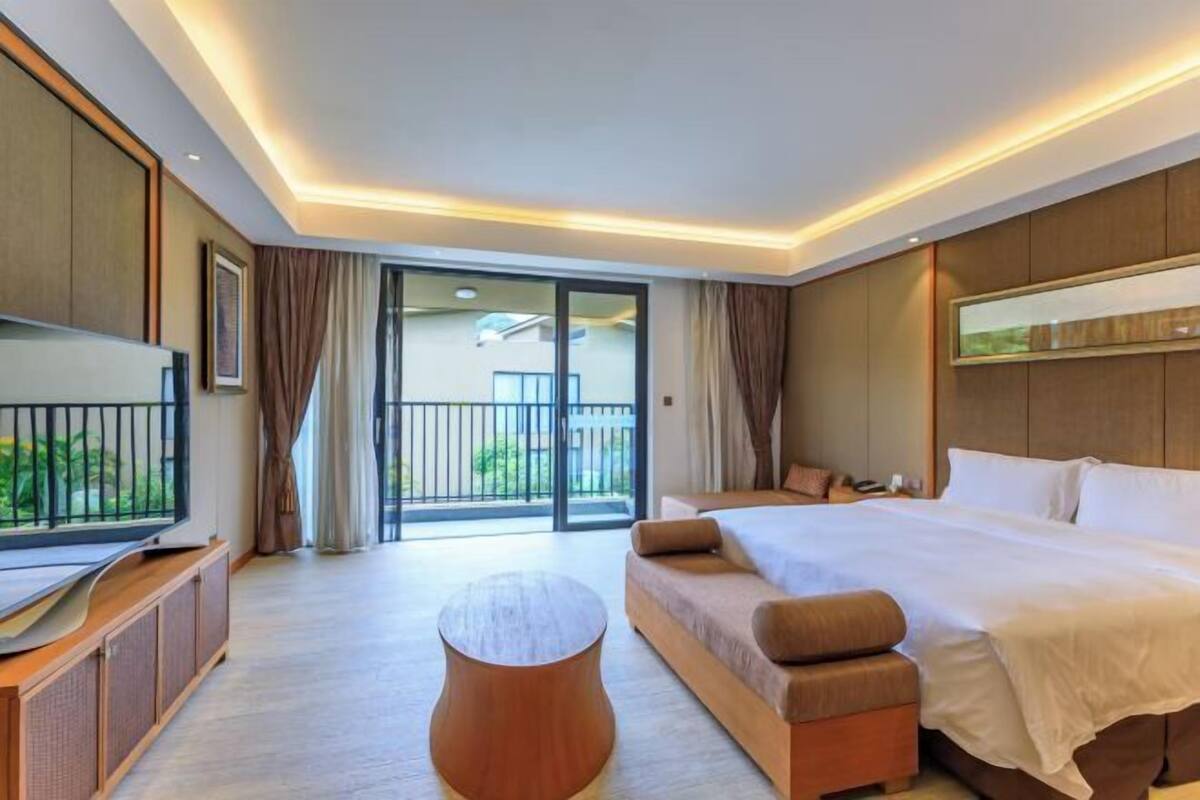 Quarto de hotel moderno com uma cama grande, televisão, uma mesa redonda pequena e uma varanda com vista para o verde.