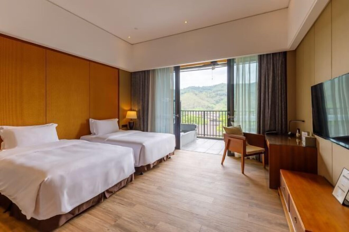 A hotel room with two beds, a desk, a chair, and a balcony with a view.