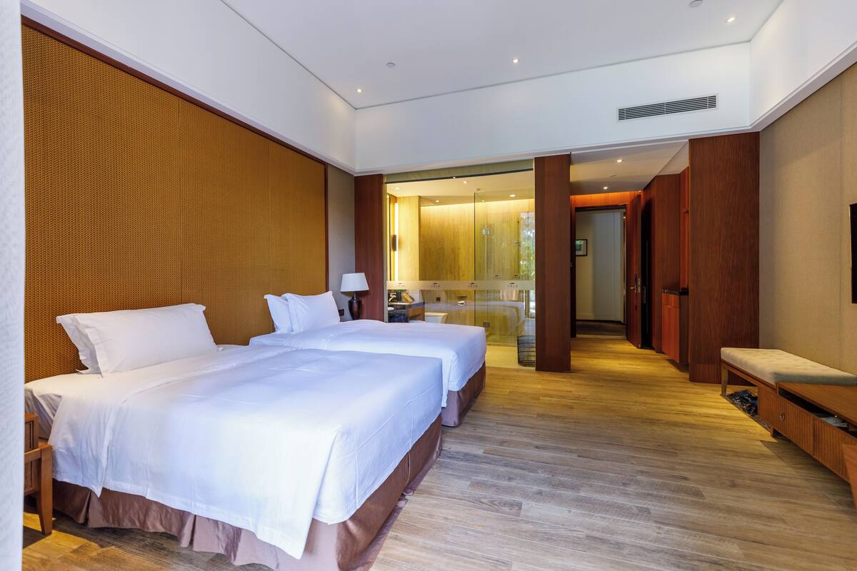 Quarto de hotel com duas camas, parede de madeira, um banco e vista para uma porta de vidro que dá para o banheiro.