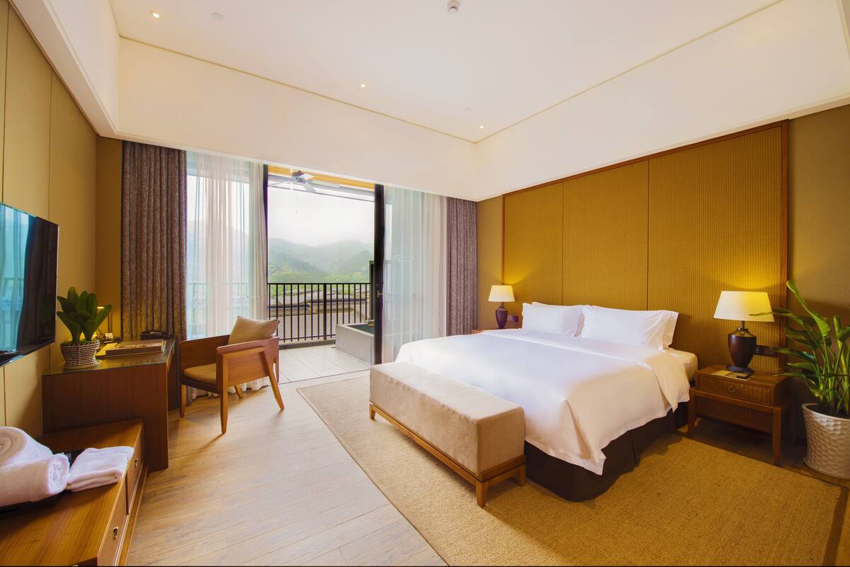 Quarto de hotel com uma cama grande, uma escrivaninha, uma cadeira, uma televisão, varanda e vista para as montanhas.