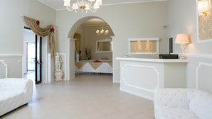 A spacious living area with a white sofa, a chandelier, and a reception desk. - Morfeo Luxury Rooms (Pozzallo)