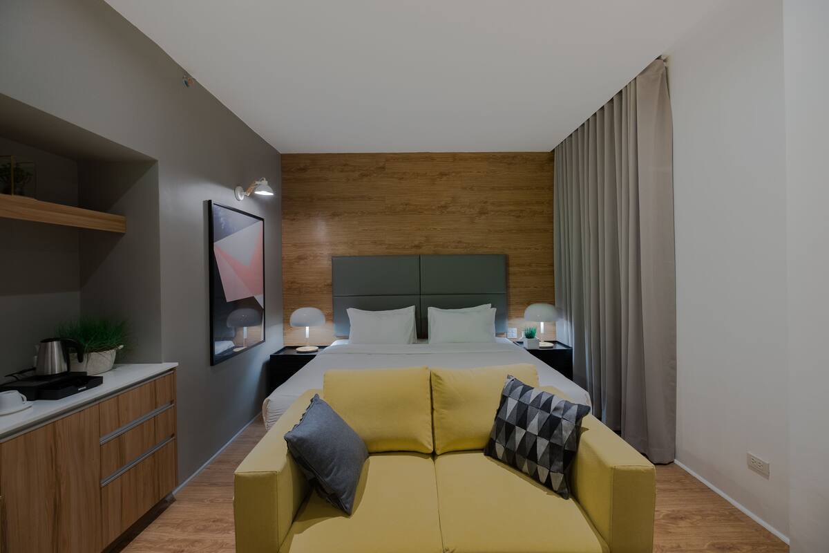 A modern hotel room with a bed, a sofa, a desk, and a nightstand.