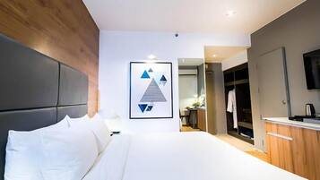 A modern hotel room with a large bed, a wooden headboard, a framed artwork on the wall, and a kitchenette area.