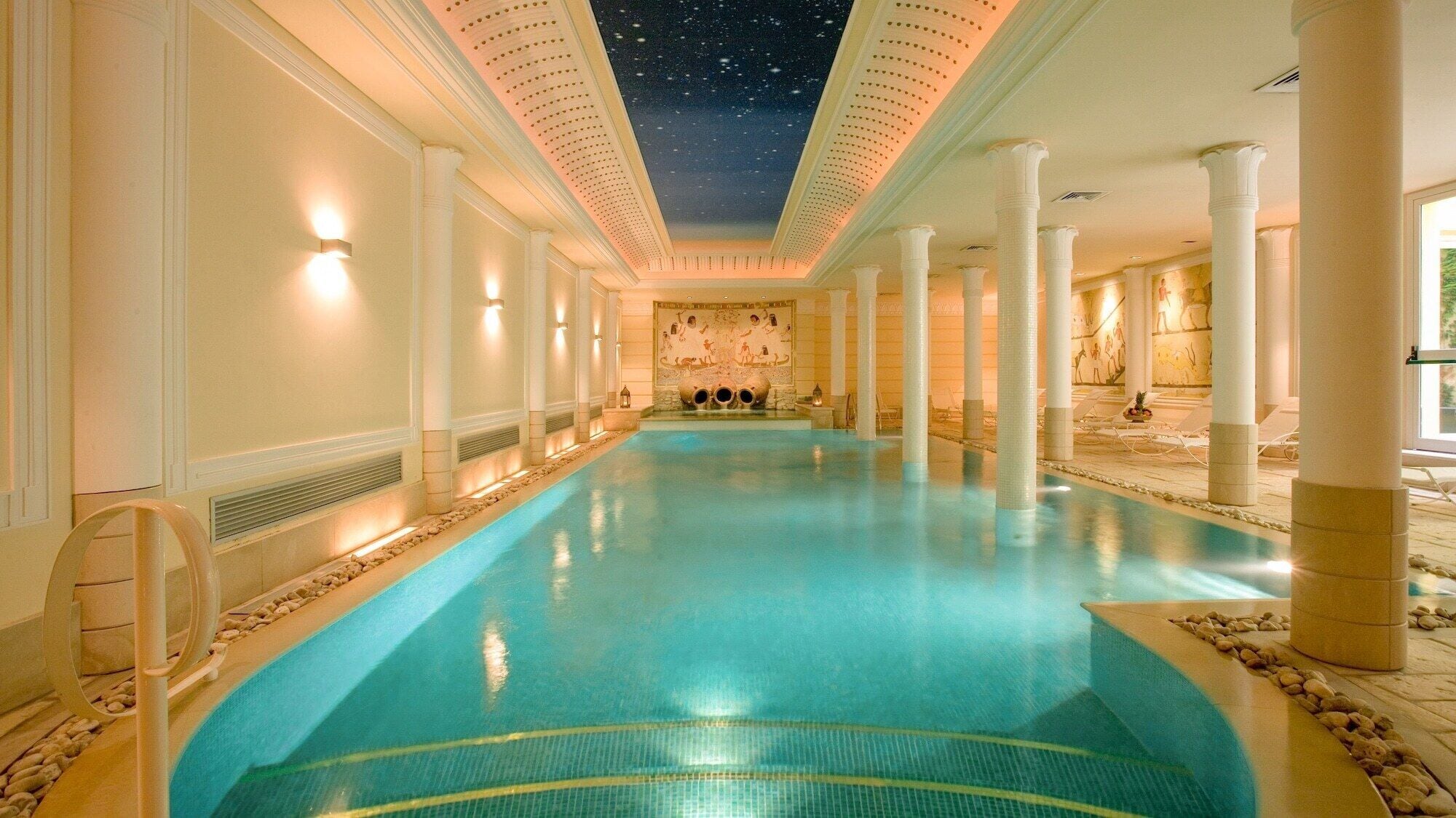 Una amplia piscina cubierta con techo estrellado, columnas blancas y pared de piedra con obras de arte.
