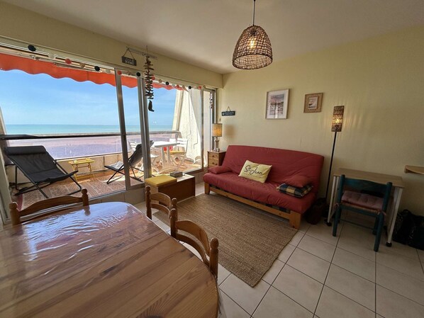 Een kamer met een eettafel, een rode bank, een houten stoel, een vloerlamp en uitzicht op het strand door grote ramen.