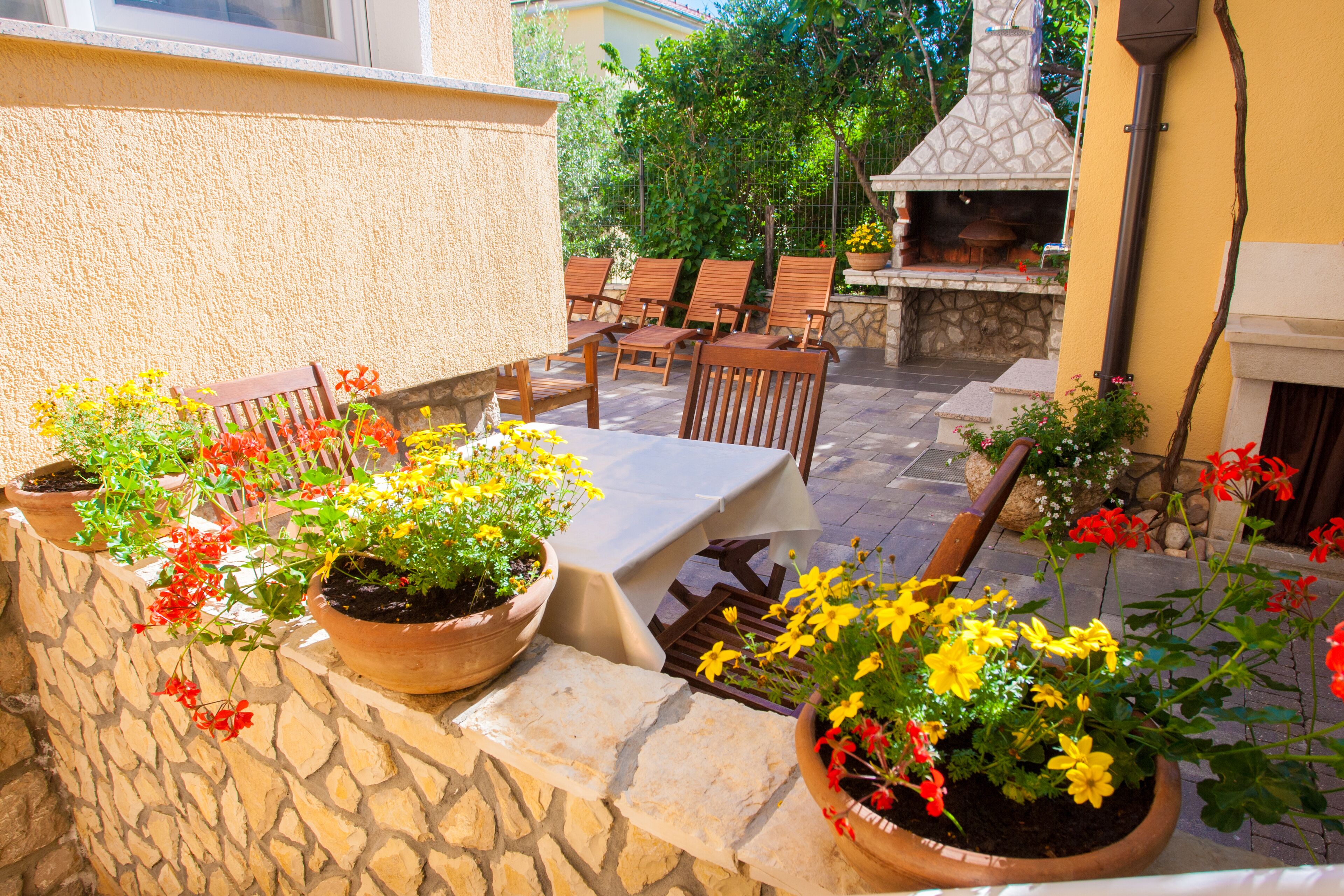 An outdoor patio with stone walls, potted plants, wooden chairs, and a table.