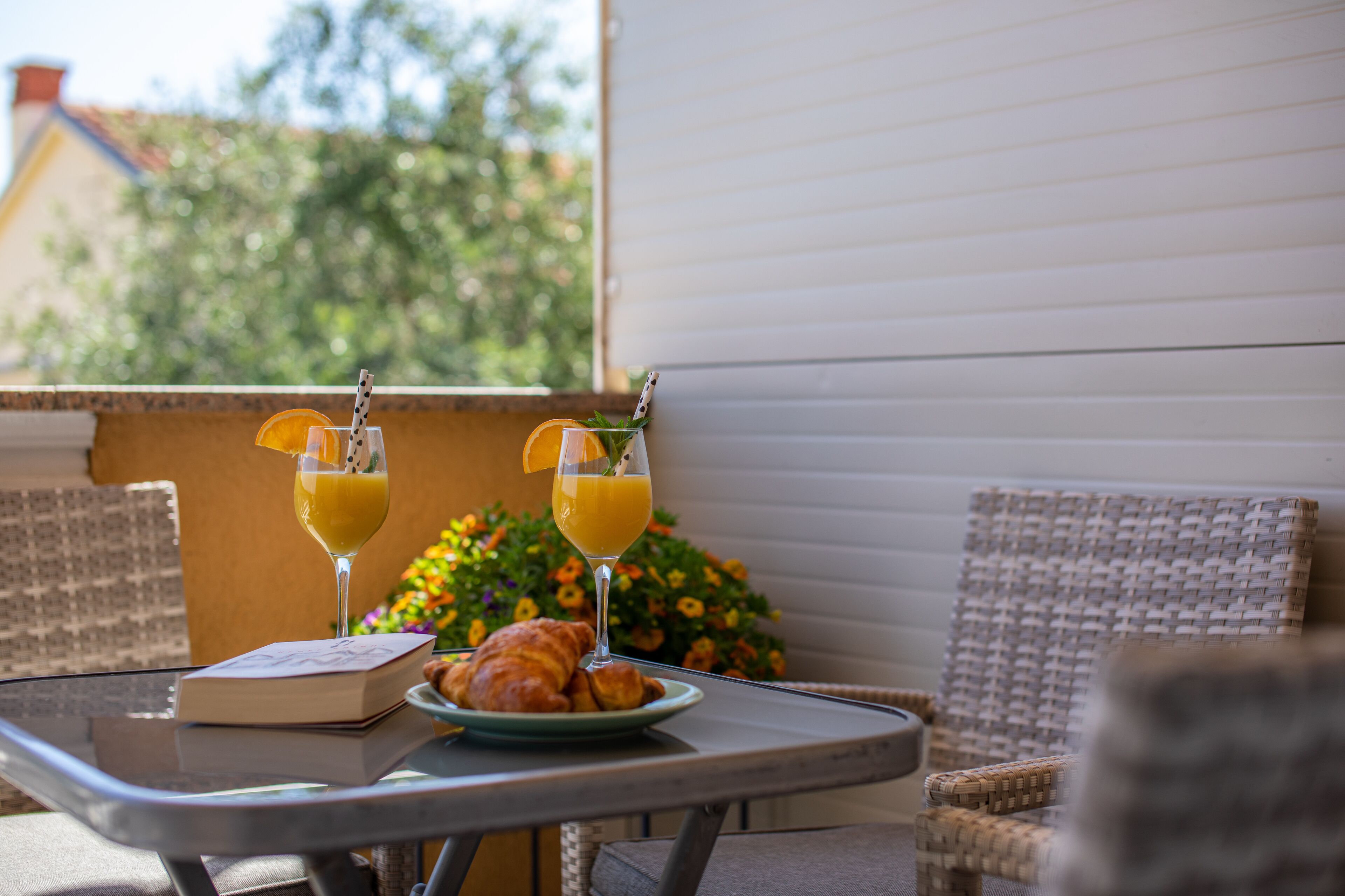 A table with a book, croissants, and two glasses of orange juice on a balcony with wicker chairs and a view of greenery.