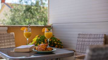 A table with a book, croissants, and two glasses of orange juice on a balcony with wicker chairs and a view of greenery.