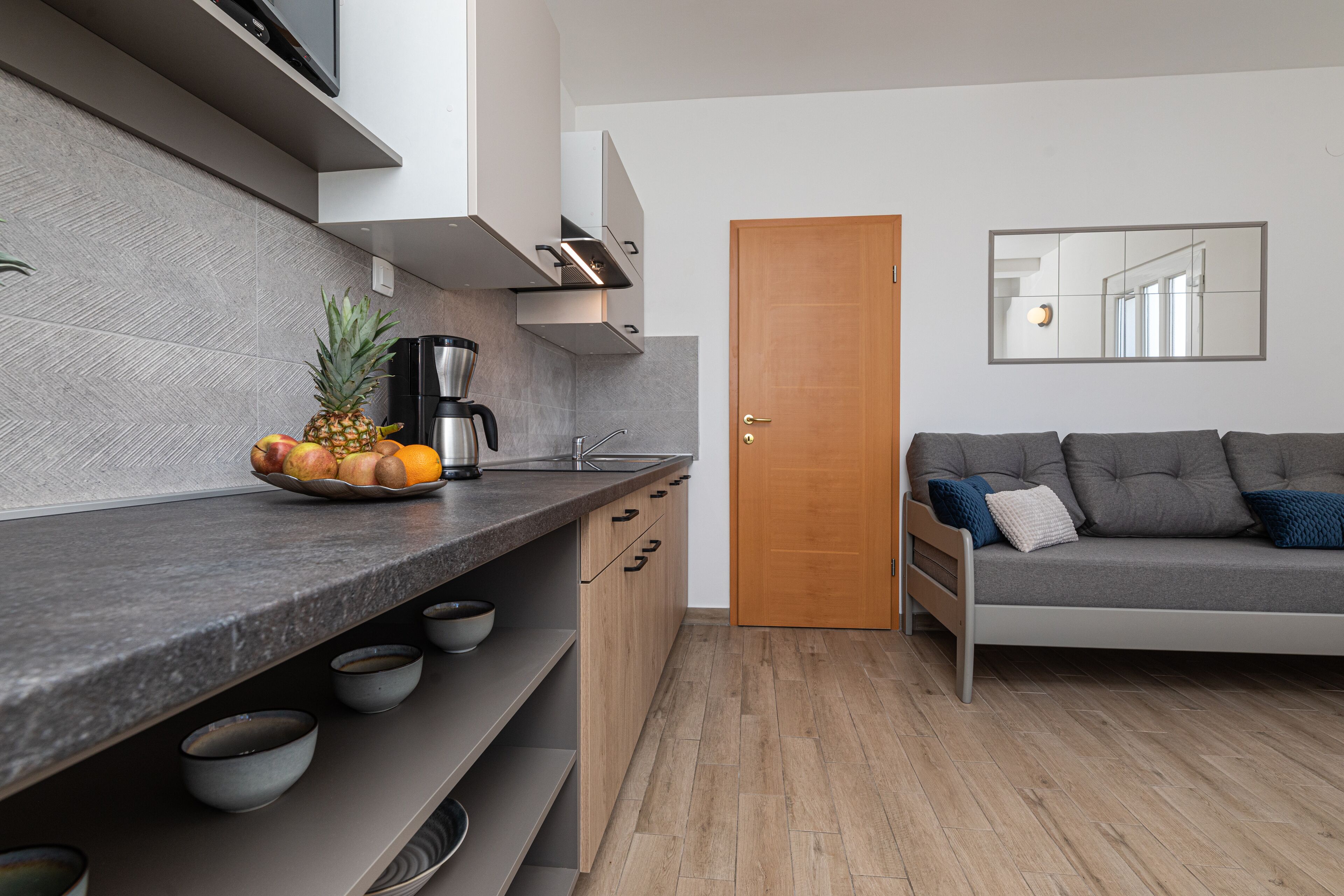 Une cuisine moderne avec un plan de travail en granit, une machine à café et un saladier rempli de fruits. On y trouve une porte en bois et un canapé gris avec des coussins.