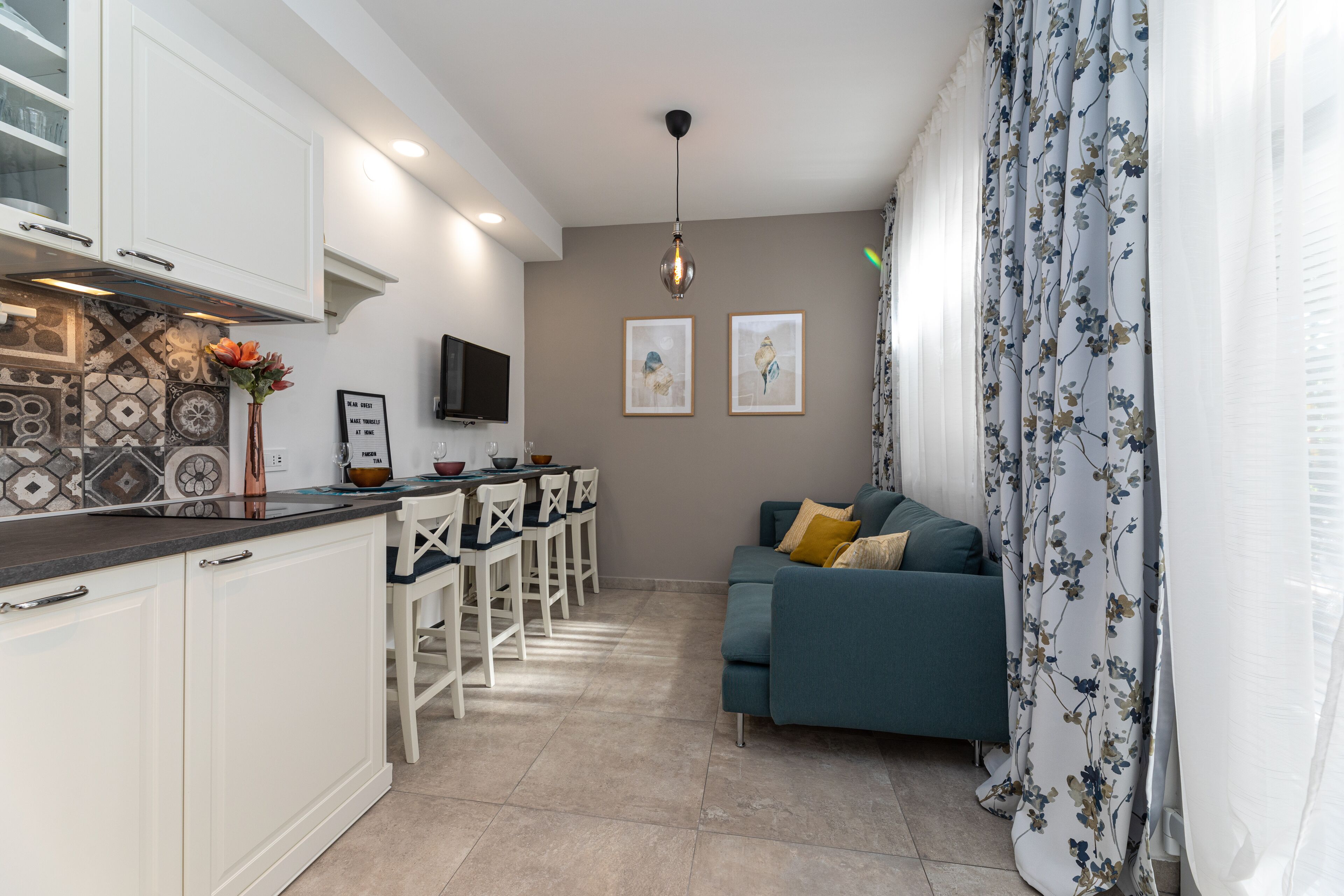 A modern kitchen with white cabinets, a dining area with a table and chairs, a teal sofa, and a TV mounted on the wall.