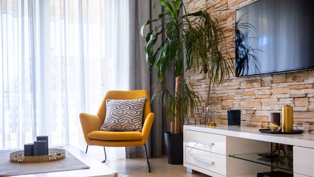 Una sala de estar moderna con un sillón amarillo, una planta grande en maceta, un televisor de pantalla plana montado en una pared de piedra y un mueble de entretenimiento blanco minimalista.
