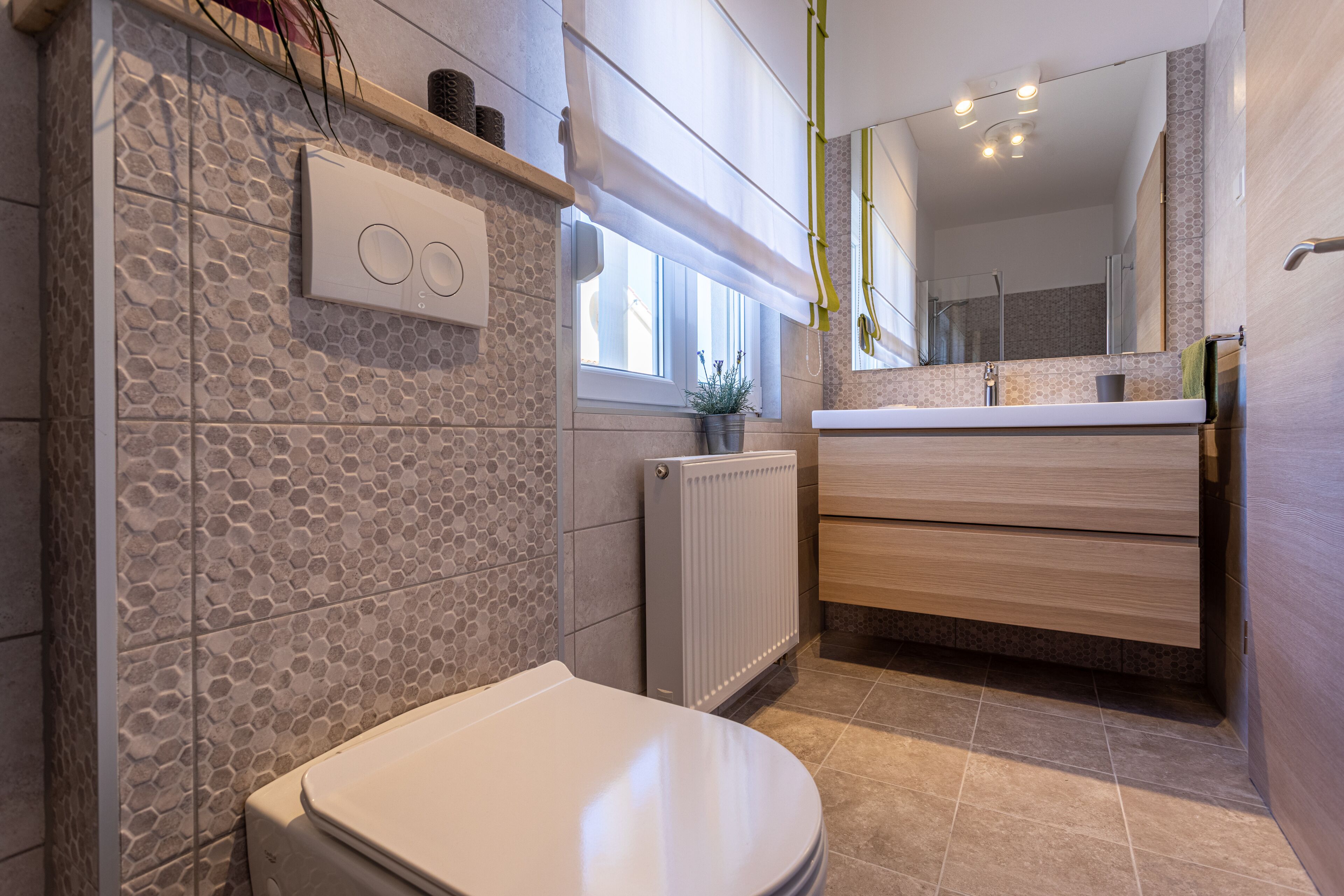 A modern bathroom with a toilet, a radiator, a window with a plant, and a wooden vanity with a mirror.