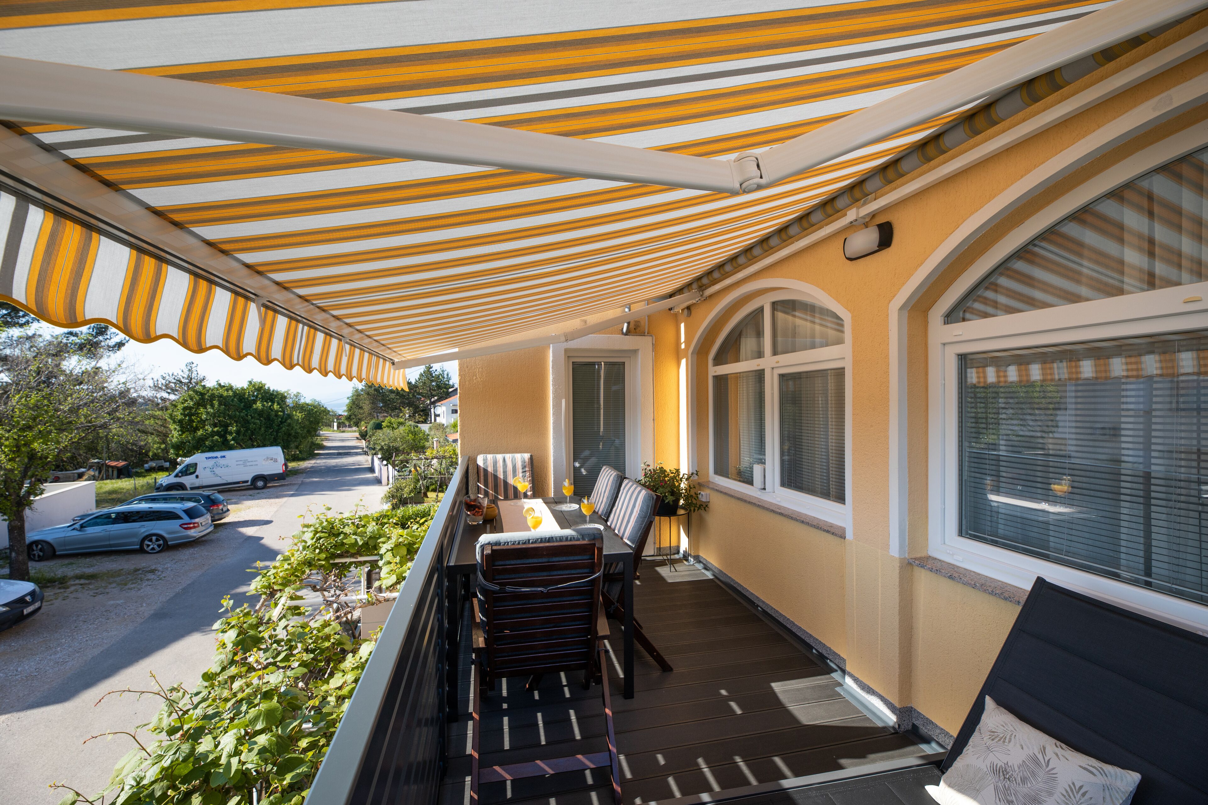 A balcony with a striped awning, a dining table with chairs, and a view of parked cars and greenery.