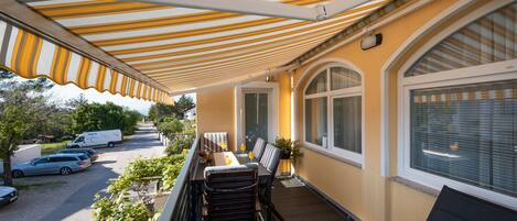 A balcony with a striped awning, a dining table with chairs, and a view of parked cars and greenery.
