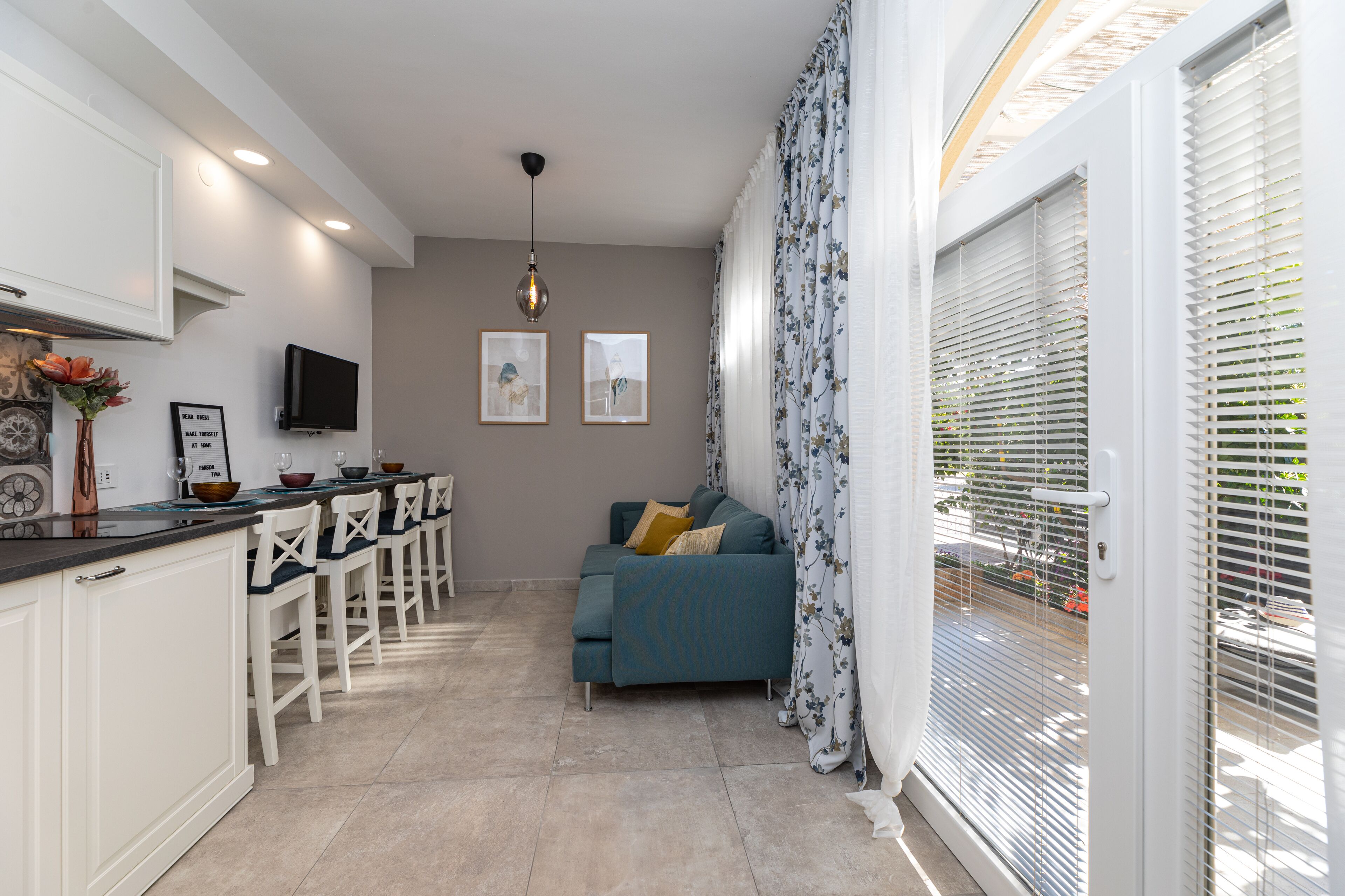 Eine moderne Küche mit weißen Schränken, ein Essbereich mit weißen Stühlen, ein blaues Sofa und ein an der Wand befestigtes Fernsehgerät.