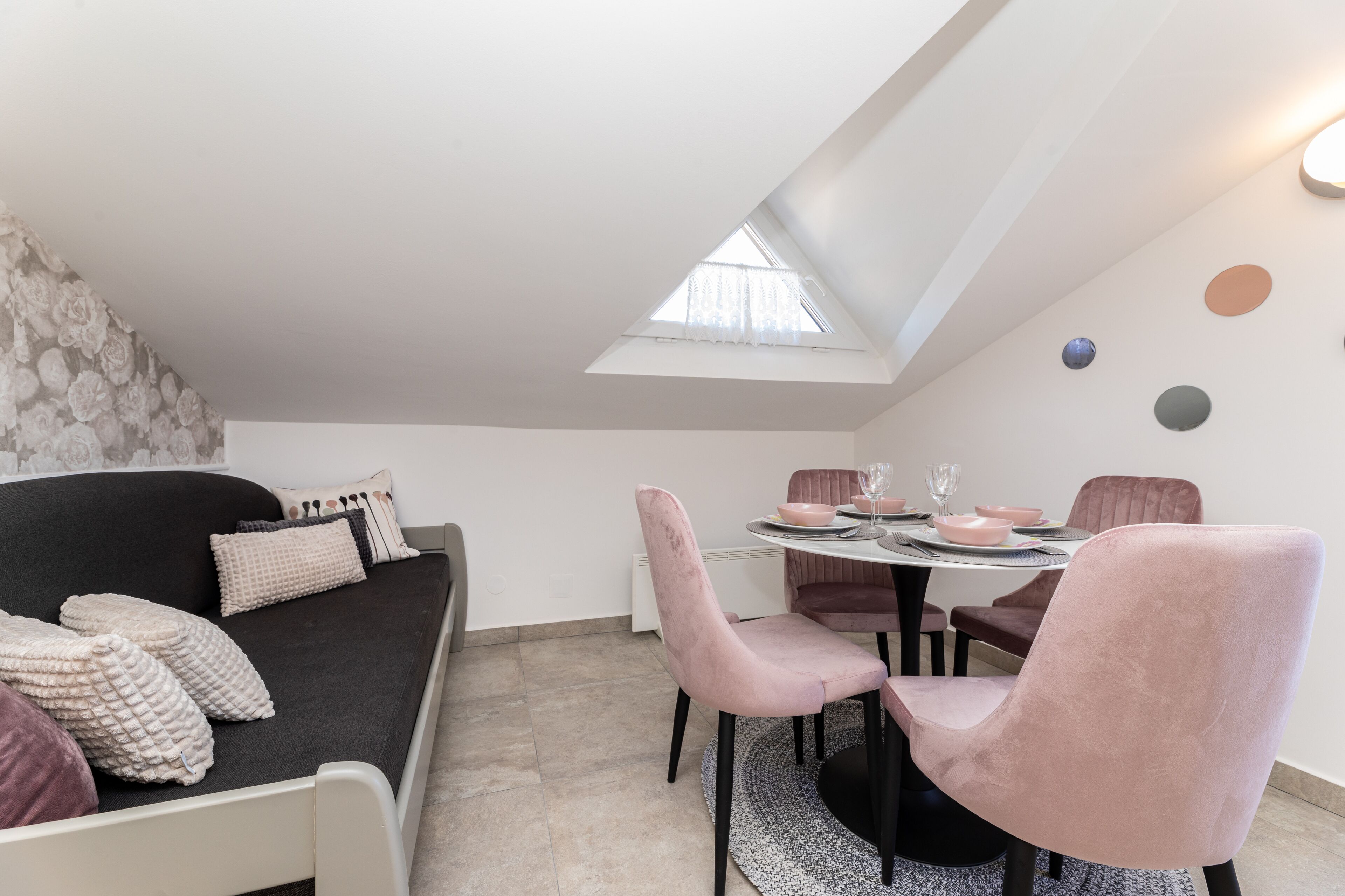 A modern loft bedroom with a skylight, a dark sofa with cushions, a dining table with pink chairs, and a patterned wall.