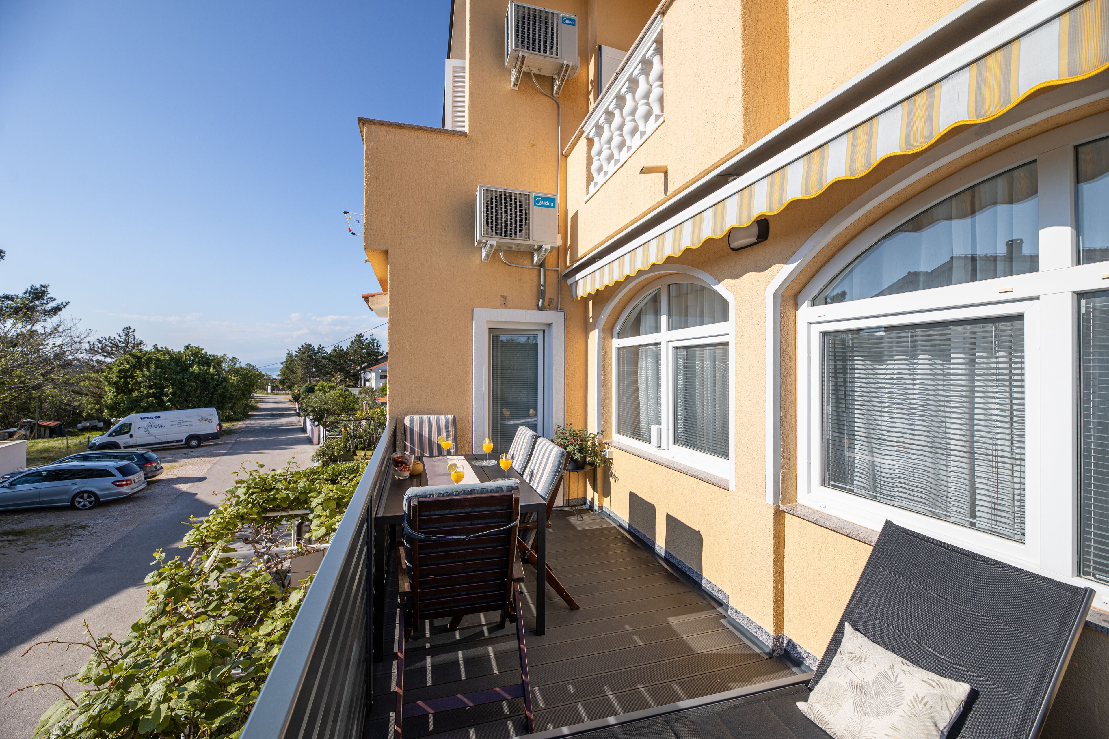 A balcony with a table and chairs, a view of the street, and a building with air conditioning units.