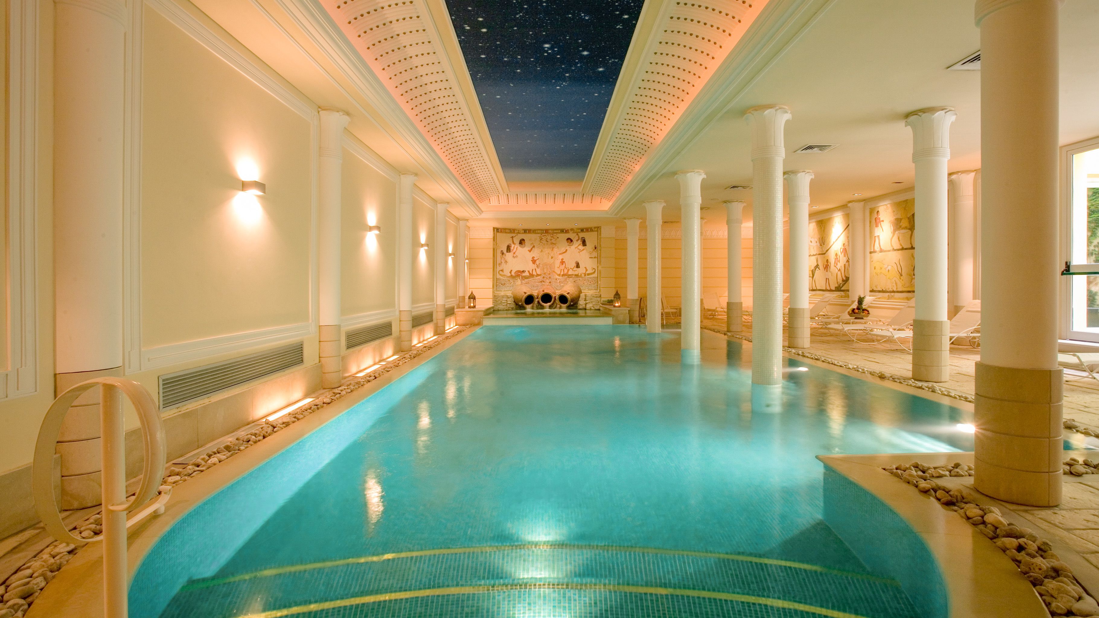 A large indoor swimming pool with a starry ceiling, white columns, and a central mural.