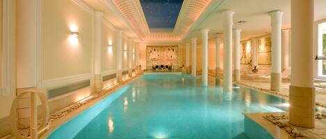 A large indoor swimming pool with a starry ceiling, white columns, and a central mural.