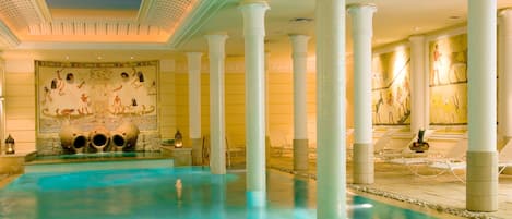 A large indoor pool with clear blue water, surrounded by white columns and Egyptian-themed wall art.