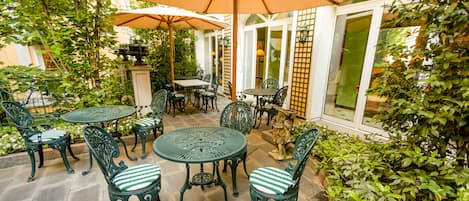 An outdoor patio with round tables and chairs, surrounded by greenery and a large umbrella.