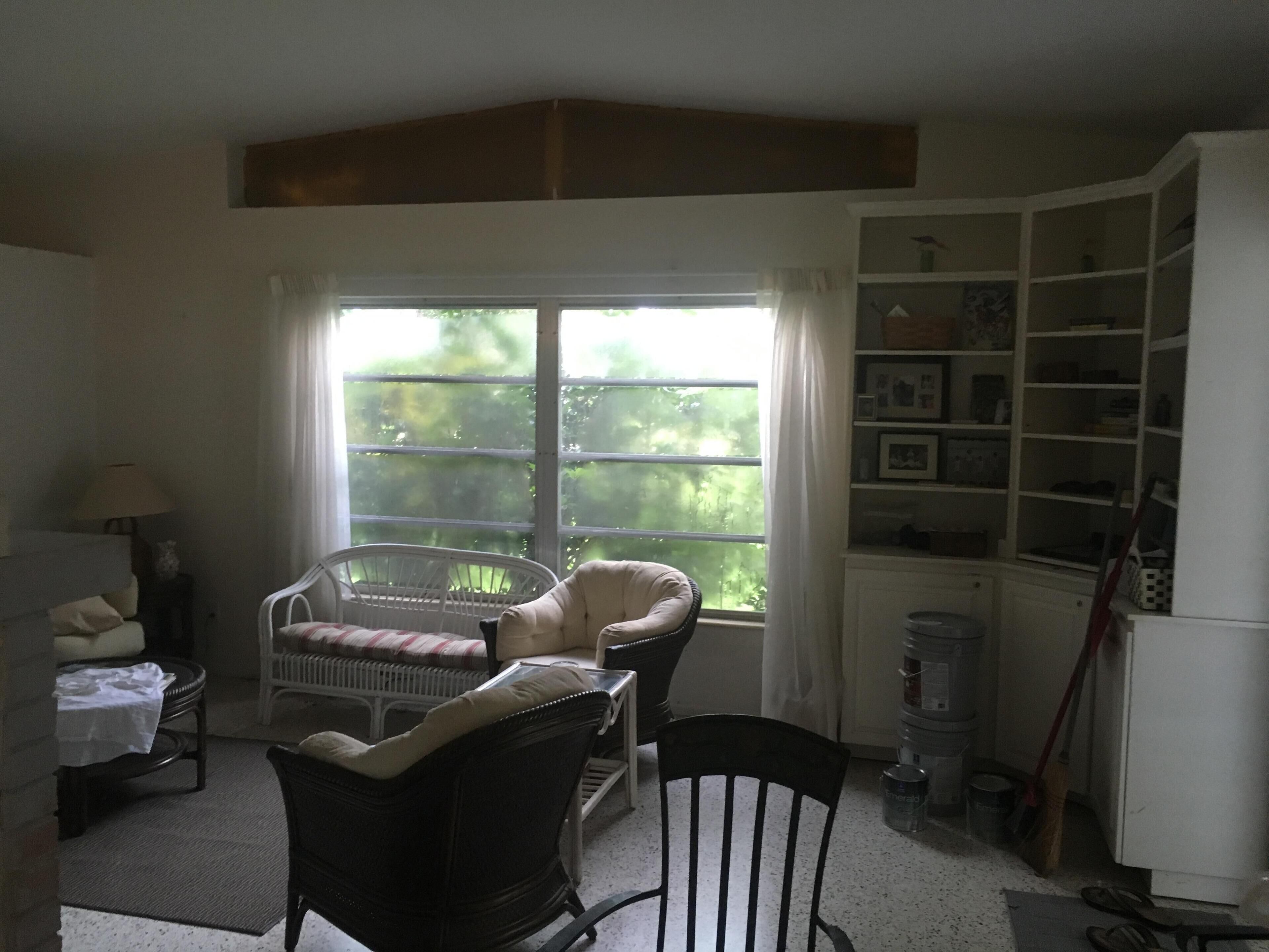 A room with a large window, a white cabinet with shelves, a wicker chair, a wooden chair, and a small table with a lamp.