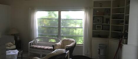 A room with a large window, a white cabinet with shelves, a wicker chair, a wooden chair, and a small table with a lamp.