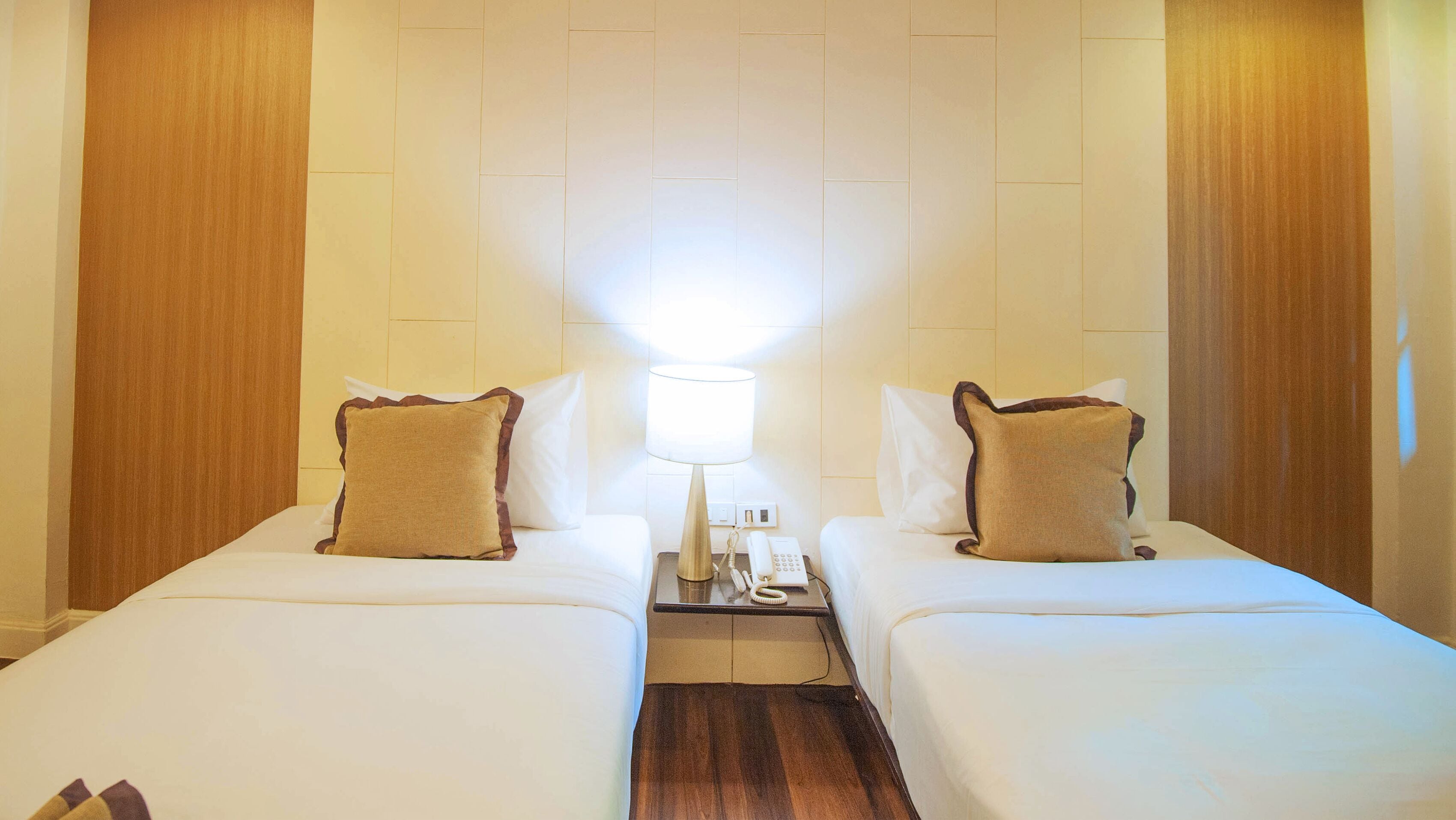 Quarto de hotel com duas camas, mesa de cabeceira com abajur e telefone, e painéis de madeira nas paredes.
