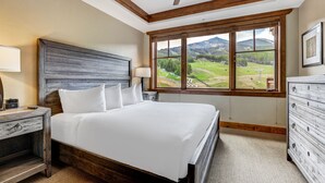 A bedroom with a large bed, wooden headboard, a nightstand with a lamp, a dresser, and a view of a green landscape through a window.