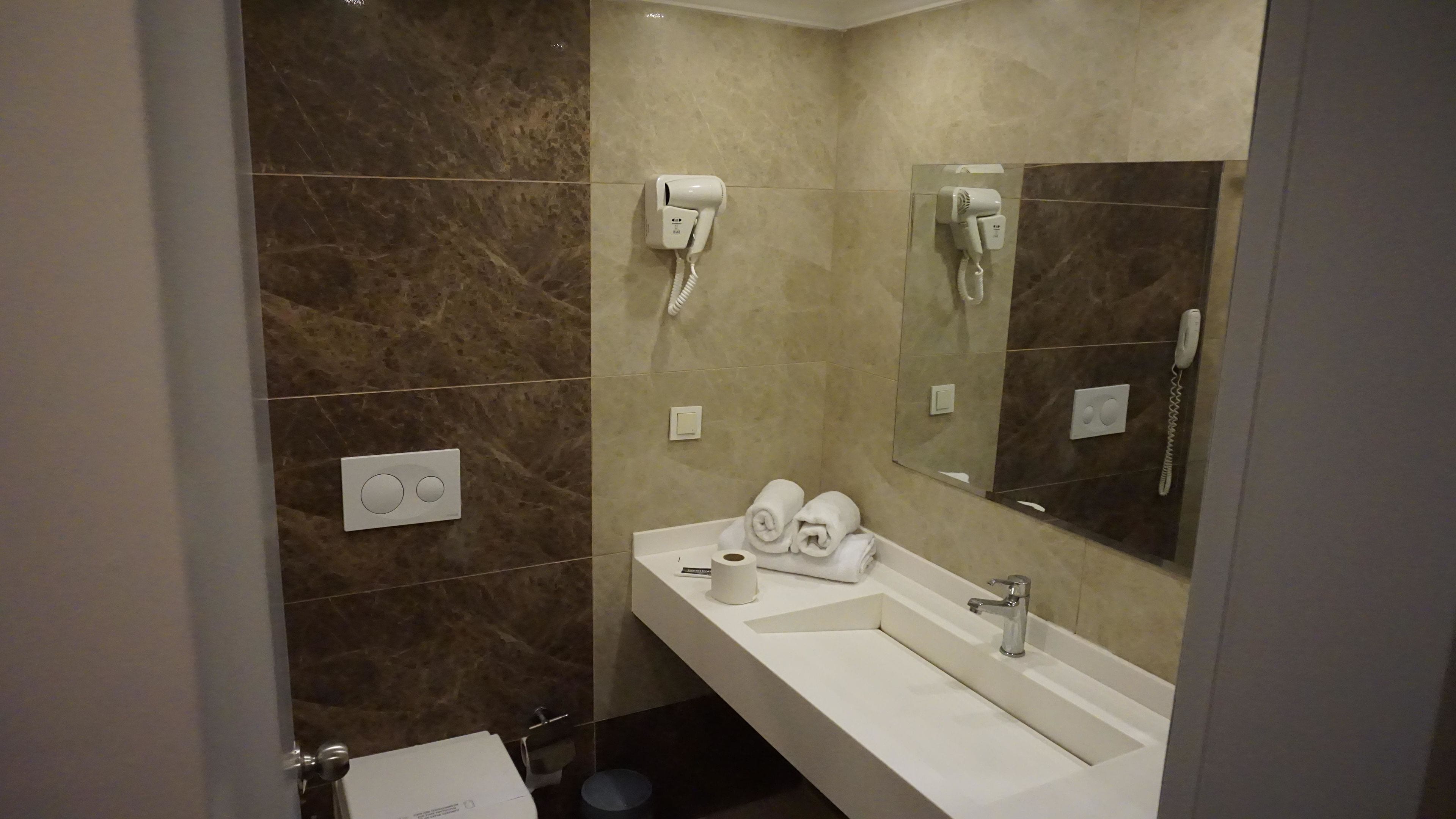 A bathroom with a marble wall, a sink with a mirror, two hair dryers mounted on the wall, and a toilet.
