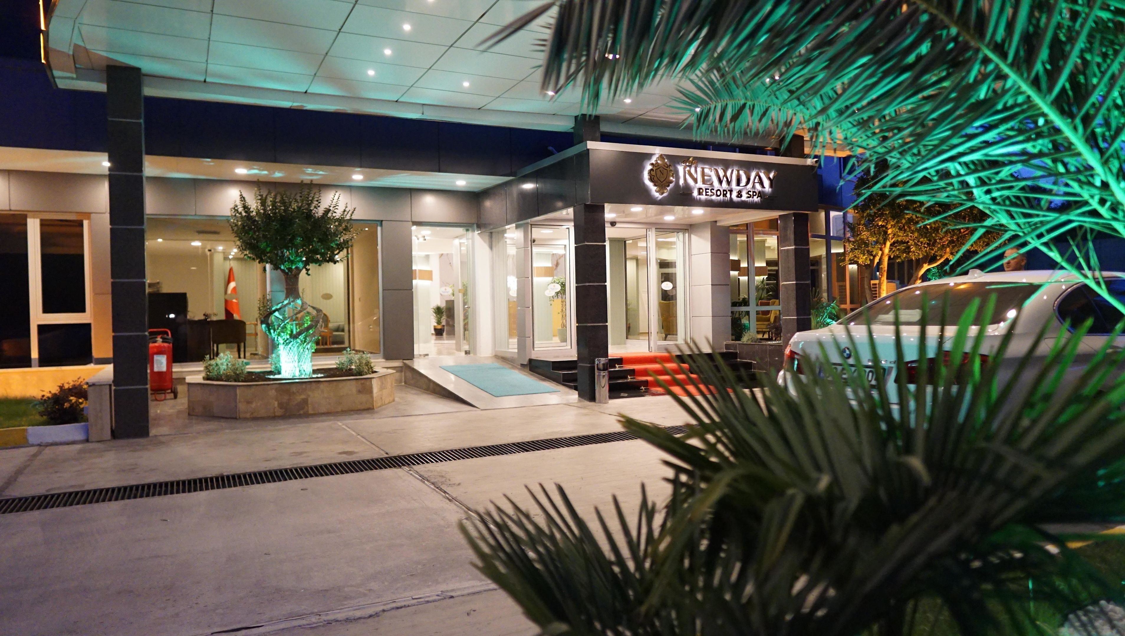 Una entrada de hotel moderna con una recepción bien iluminada, un jardín ajardinado con un árbol y un coche BMW estacionado afuera.