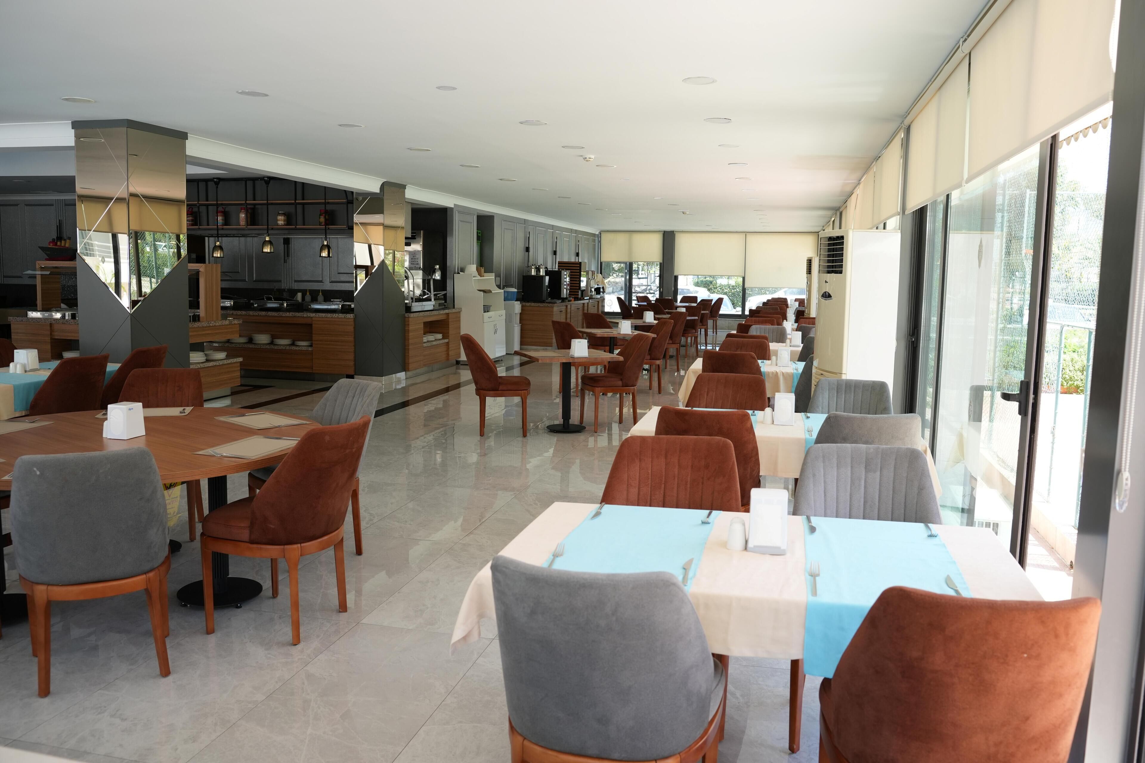 A spacious dining area with multiple tables and chairs, a kitchen in the background, and large windows providing natural light.