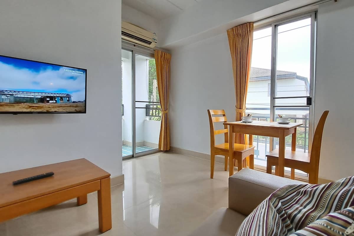 A modern hotel room with a balcony, a dining table with chairs, a sofa, and a television mounted on the wall.