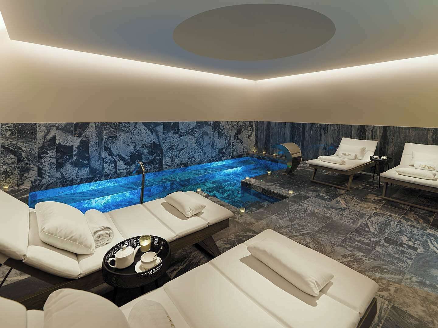 A spa room with a hot tub, two lounge chairs, a massage table, and a wall with a water feature.