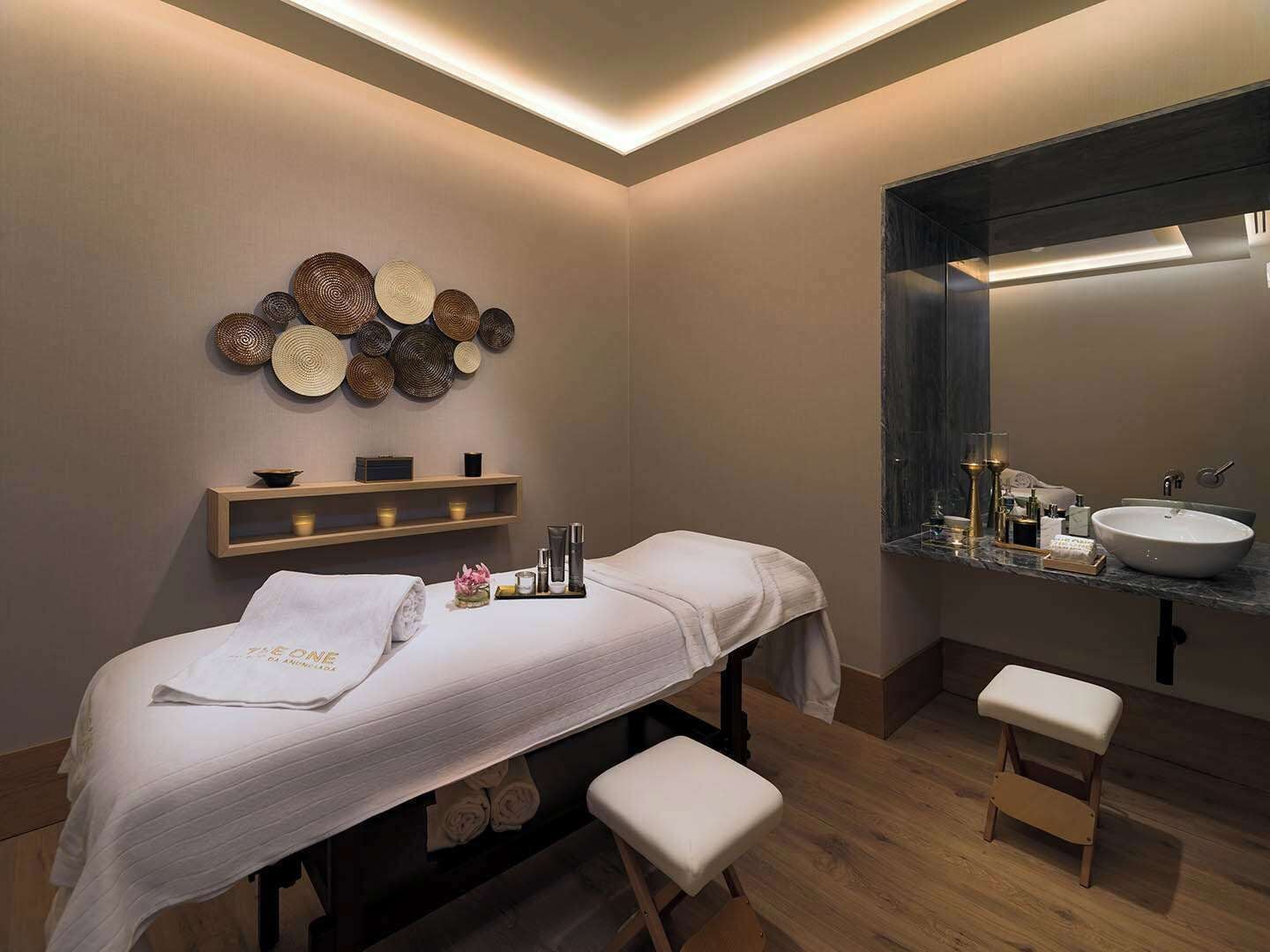 A modern spa room with a massage table, a wooden shelf with candles, and a large mirror above a sink.