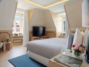 A modern hotel room with a large bed, a flat-screen TV, a bedside table with a vase of flowers, and a view of the cityscape through the window.