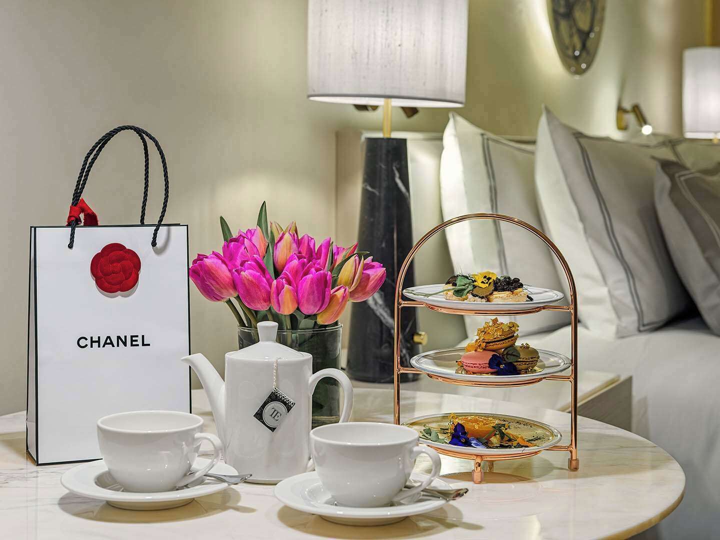 Quarto de hotel com jogo de chá, uma bolsa Chanel, um vaso de tulipas e um suporte escalonado para sobremesas.