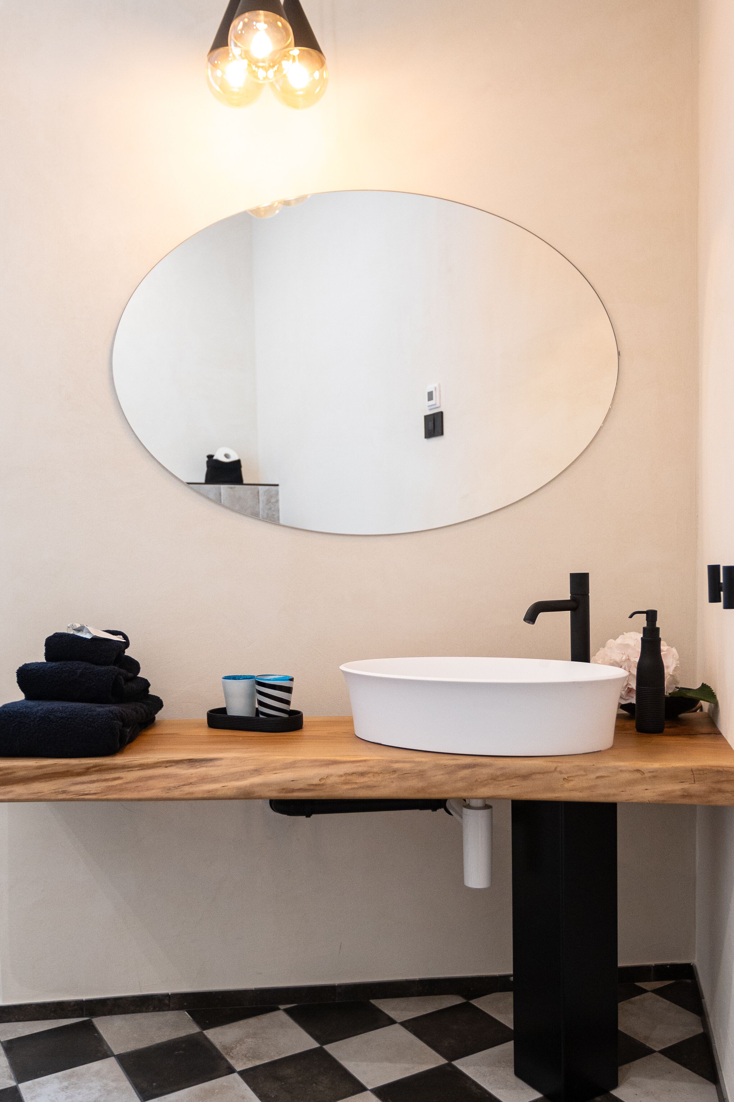Un baño con un espejo redondo, un lavabo blanco y un grifo negro. Hay toallas y una botella en la encimera.