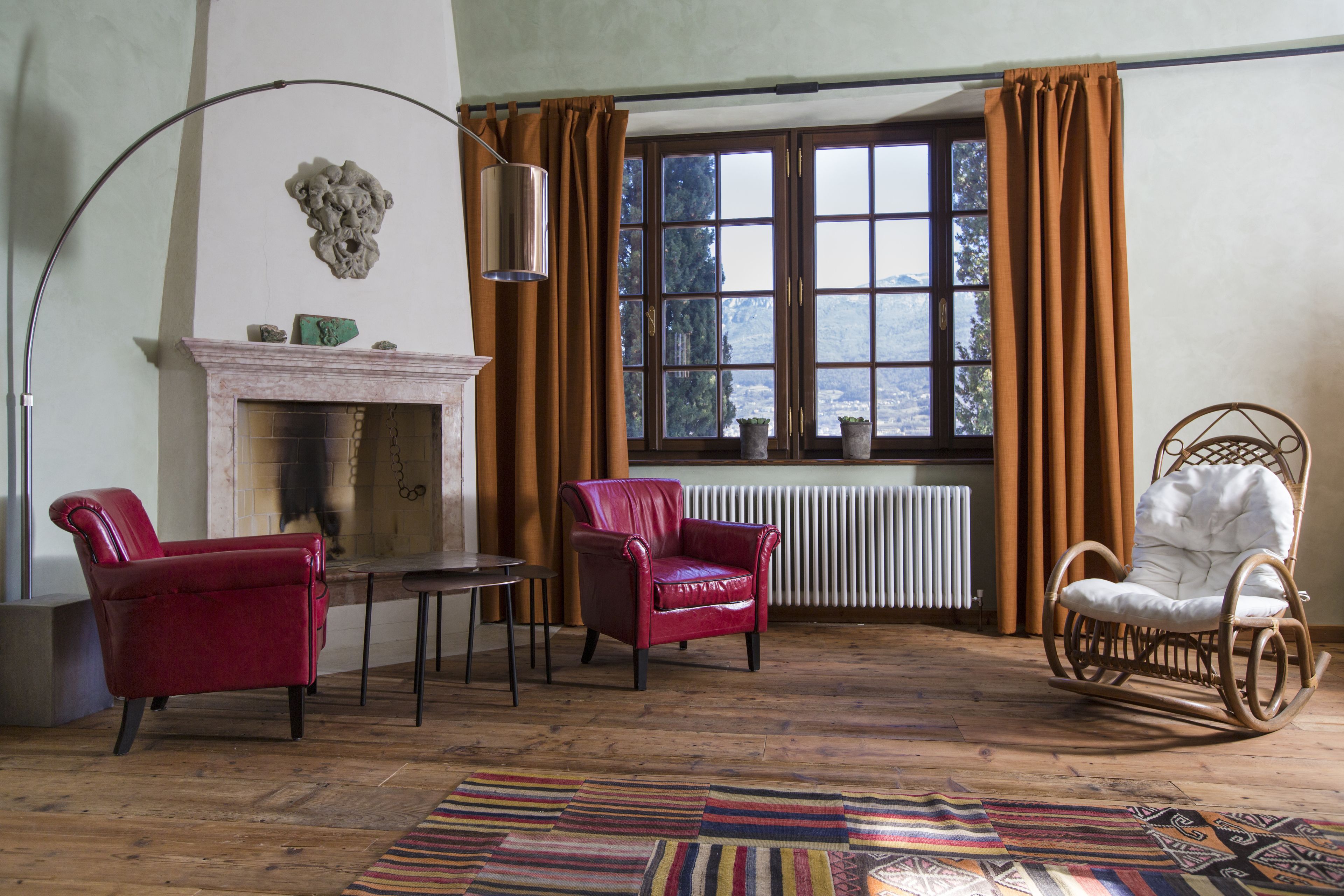 Ein Zimmer mit Kamin, zwei Ledersesseln, einem Korbsessel und einem Teppich mit Muster. Ein großes Fenster mit Vorhängen.