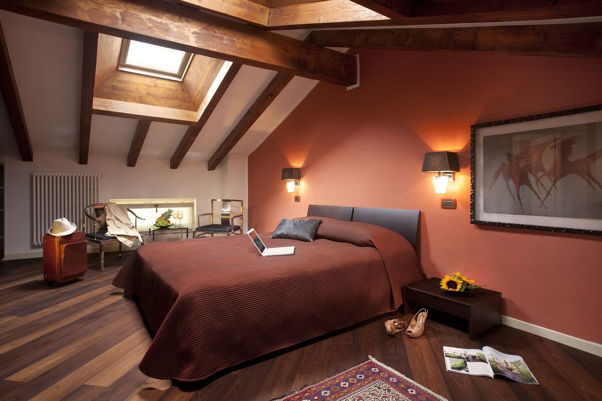 A bedroom with a sloped ceiling, wooden beams, a bed with a laptop, a desk with a chair, and a wall-mounted lamp.