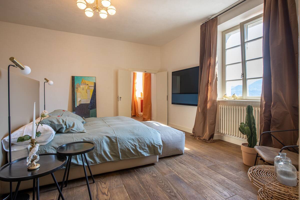 A bedroom with a bed, bedside tables, a potted cactus, a wall-mounted TV, and a window with curtains.