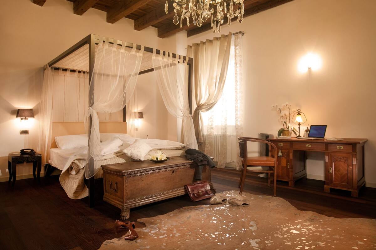 A four-poster bed with a canopy, a wooden desk with a lamp, and a leather bag on the floor.