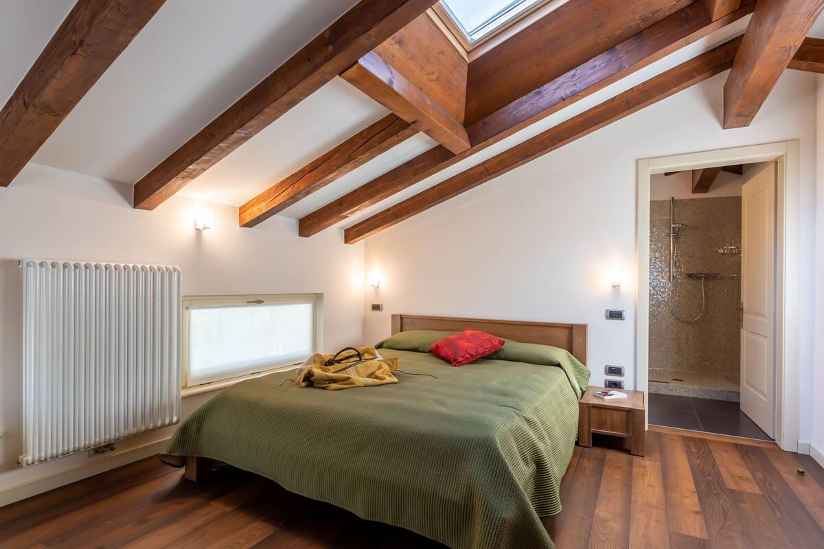 A bedroom with a sloped ceiling, wooden beams, a bed with green bedding, a red pillow, a nightstand, and a walk-in shower.
