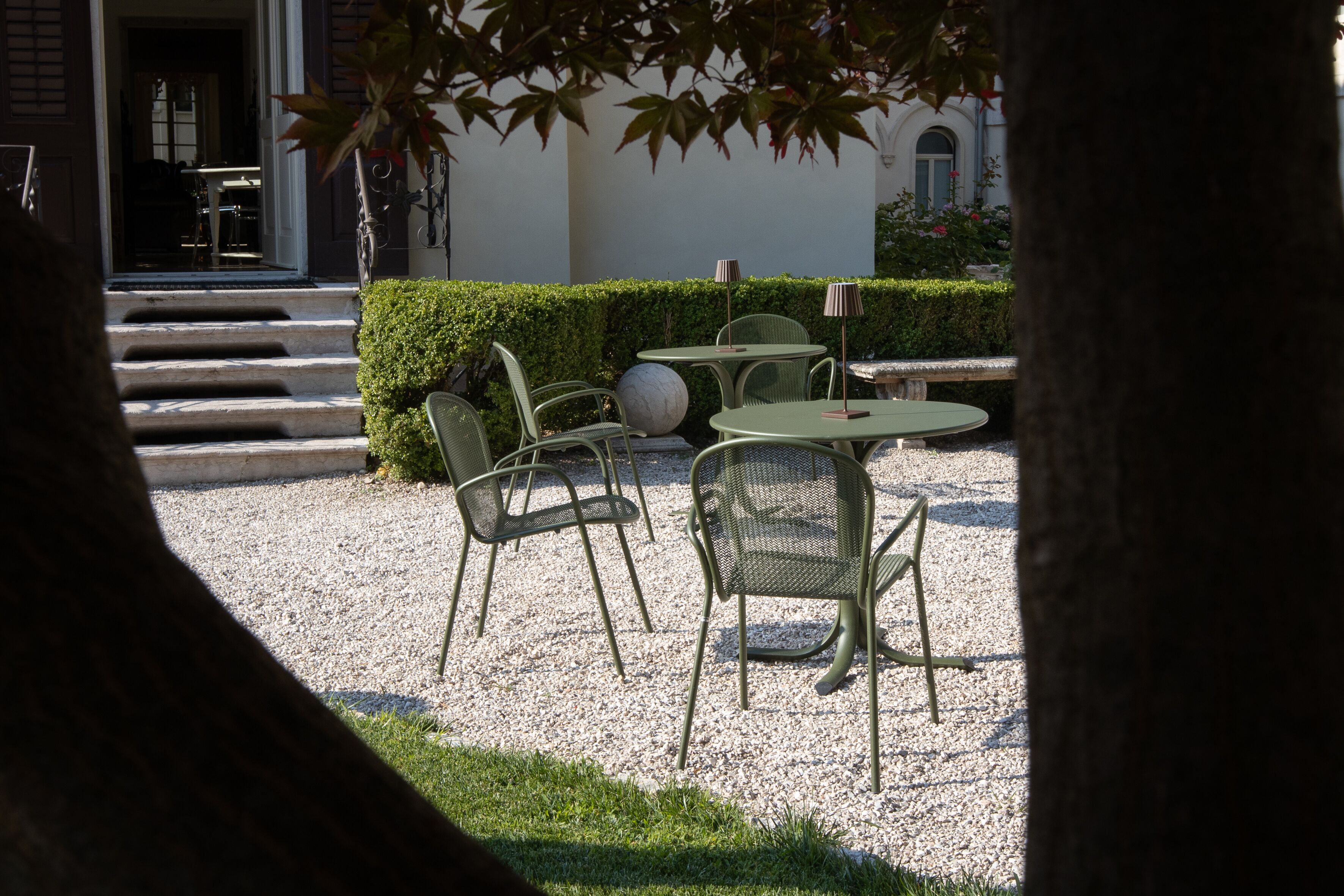 A courtyard with outdoor seating, including round tables and chairs, surrounded by a gravel area and a hedge.